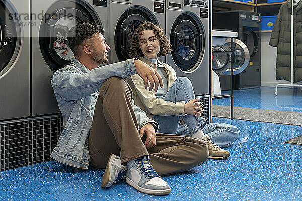 Smiling young customers sitting together leaning on washing machines at laundromat