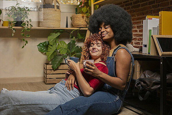 Young woman with eyes closed leaning on friend sitting on floor having coffee at home