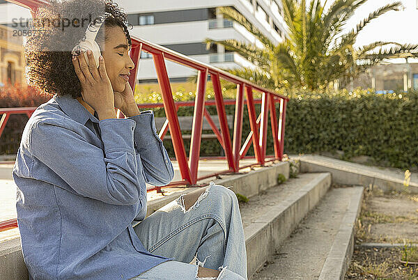 Carefree woman listening music through wireless headphones and sitting on staircase at park