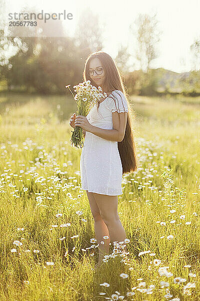 Smiling young woman holding bunch of chamomile standing in field