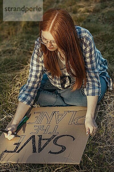 Focused redhead girl making protest banner sitting on grass at park