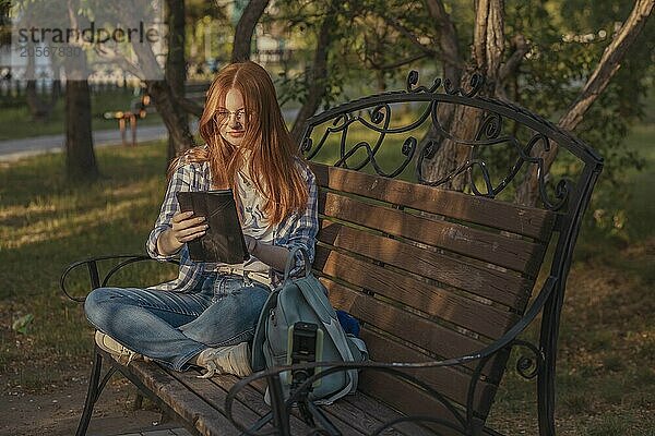 Teenage freelancer using tablet PC sitting cross-legged on bench and vlogging through smart phone at park