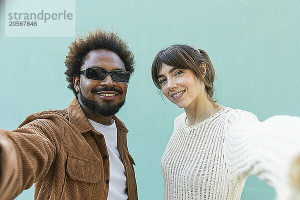 Happy multiracial couple taking selfie in front of blue wall