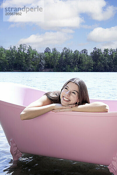 Cheerful girl relaxing inside pink bathtub under cloudy sky in lake