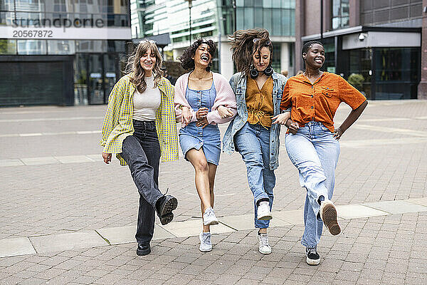 Happy female friends dancing arm in arm on footpath in city