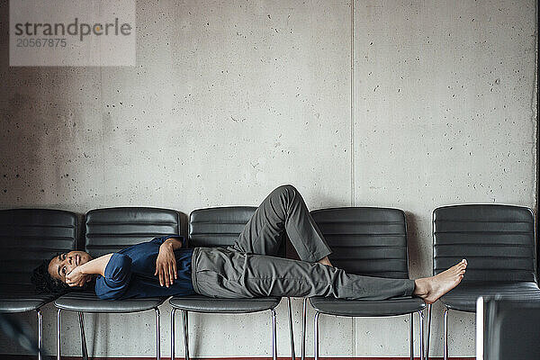 Young businesswoman lying on chairs talking through mobile phone in front of wall at office lobby