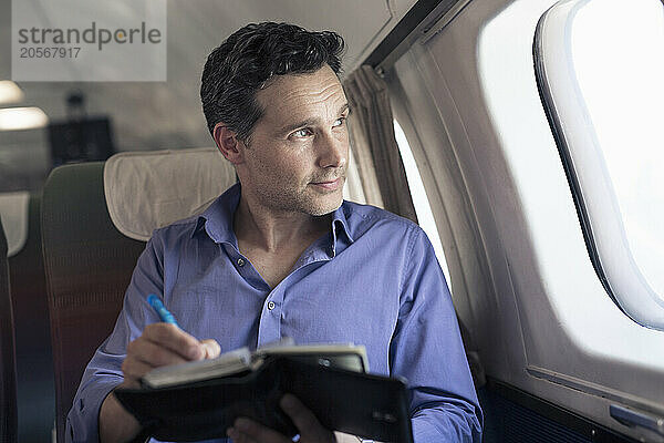 Confident mature businessman with diary looking out through airplane window