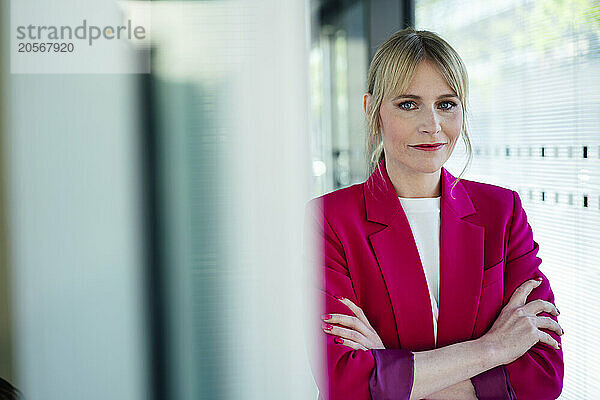 Confident blond businesswoman wearing pink blazer with arms crossed at office