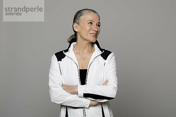 Smiling senior woman standing with arms crossed looking away against gray background