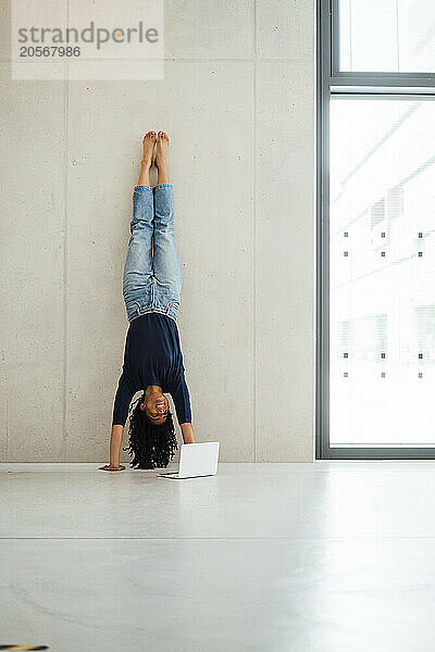 Businesswoman leaning on wall doing handstand near laptop at office lobby