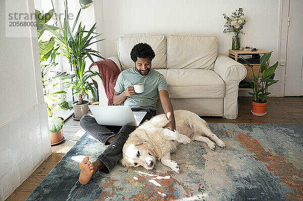 Young freelancer with Golden Retriever sitting on carpet in living room at home