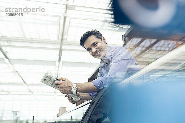 Smiling businessman with newspaper leaning on railing at airport terminal