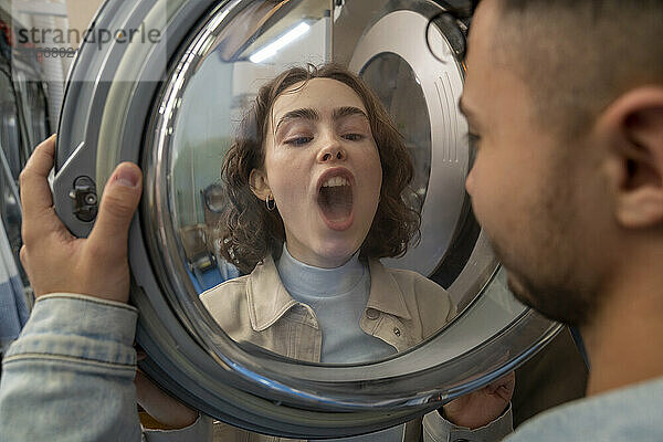 Young man looking at girlfriend breathing on washing machine glass in laundromat