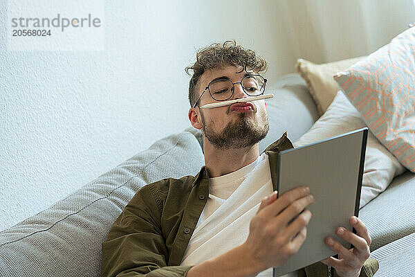 Playful young man with digitized pen on puckering lips using tablet PC at home