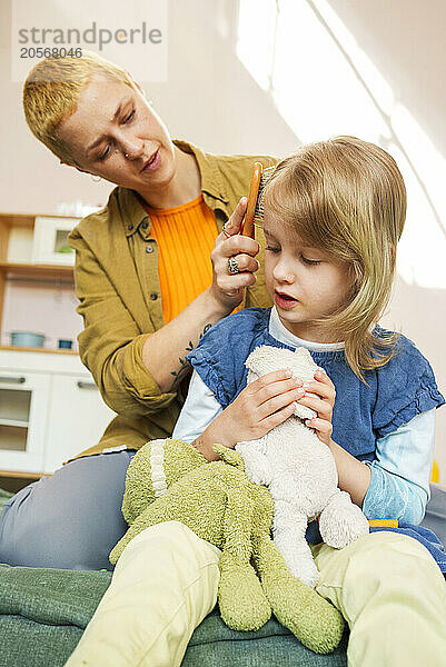 Mother combing hair of daughter sitting at home