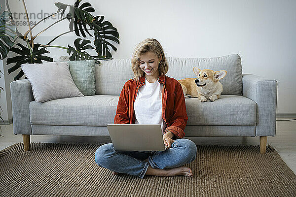 Smiling freelancer working on laptop with dog in living room at home