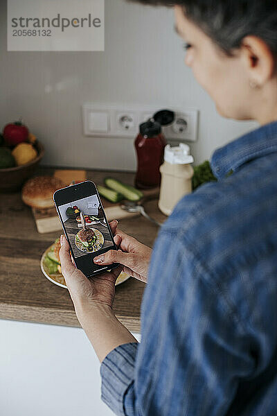 Woman taking photo of burger through smart phone in kitchen