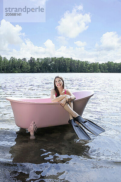 Girl with diving flipper sitting inside pink color bathtub in lake under cloudy sky