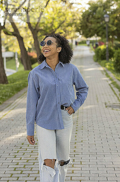 Happy young woman wearing sunglasses and walking with hand in pocket on footpath at park