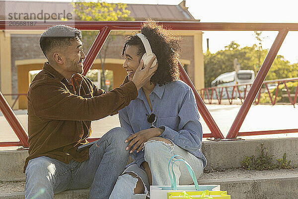 Happy young man with girlfriend wearing wireless headphones and sitting on staircase at park