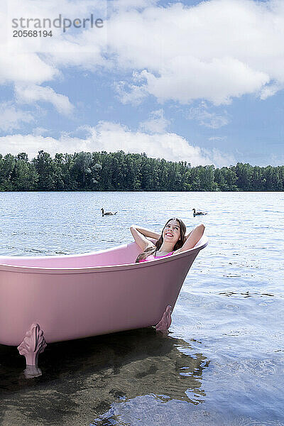 Happy girl lying and relaxing inside pink bathtub under cloudy sky in lake