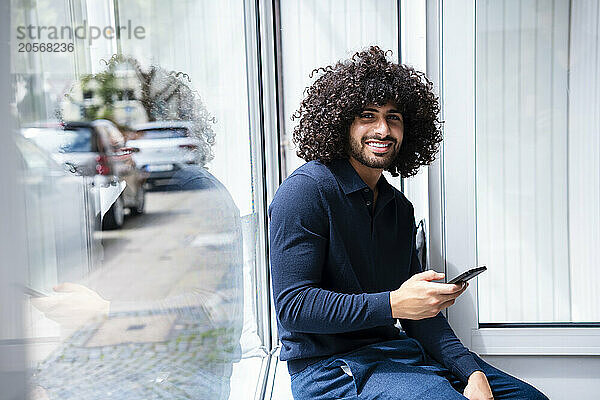 Smiling businessman with mobile phone sitting on window sill at office