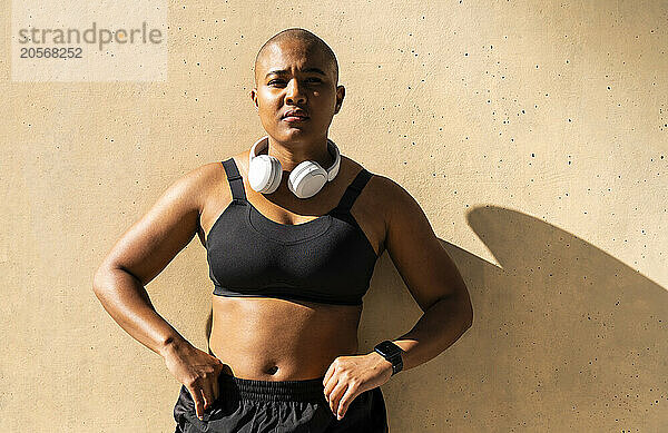 Confident muscular woman standing with arms akimbo in front of wall on sunny day