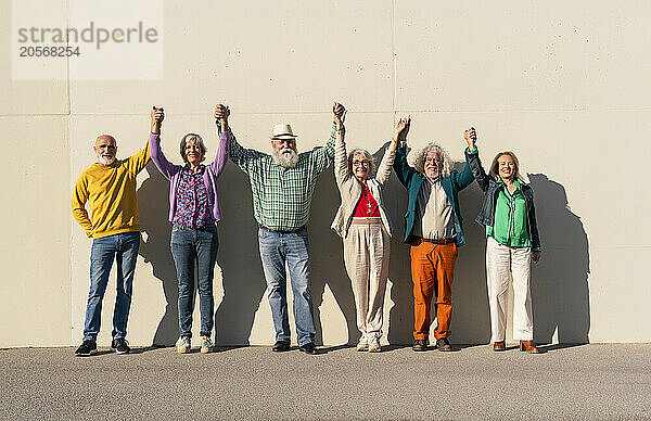 Retired women and men holding hands standing with arms raised in front of wall on sunny day