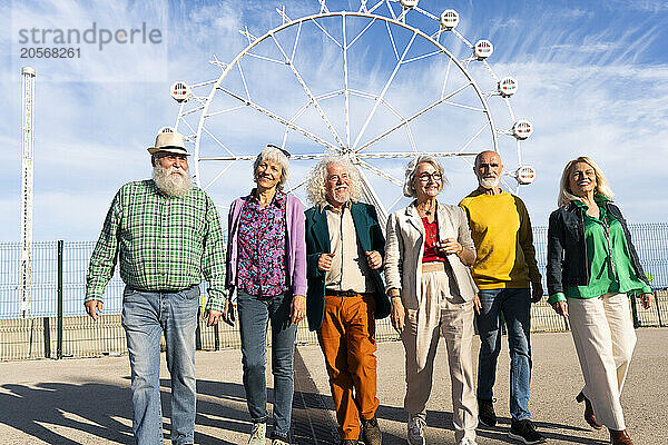 Retired men and women walking side by side in front of Ferris wheel on sunny day