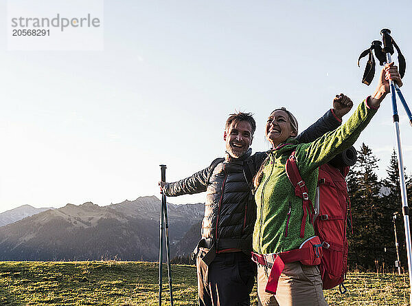 Carefree couple holding hiking poles and arms outstretched standing under clear sky in Tyrol