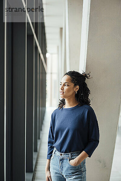 Thoughtful young businesswoman standing with hands in pockets looking out through window at office