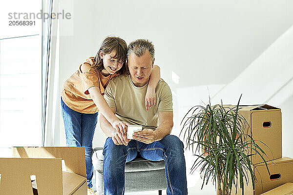 Father and daughter using smart phone amidst cardboard boxes in new house