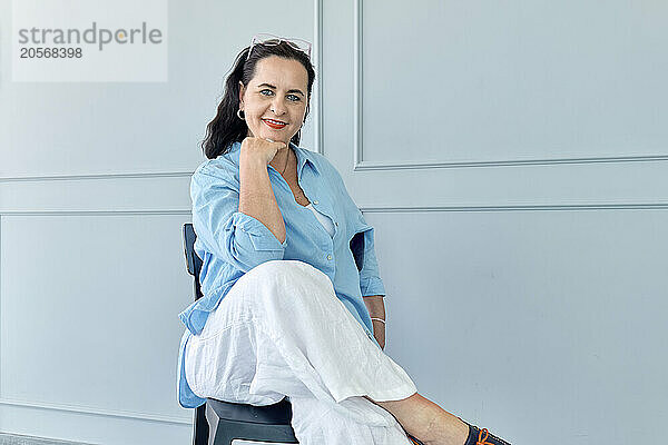 Smiling businesswoman sitting with hand on chin near wall at office