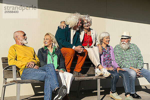 Happy retired friends sitting together on bench in front of wall