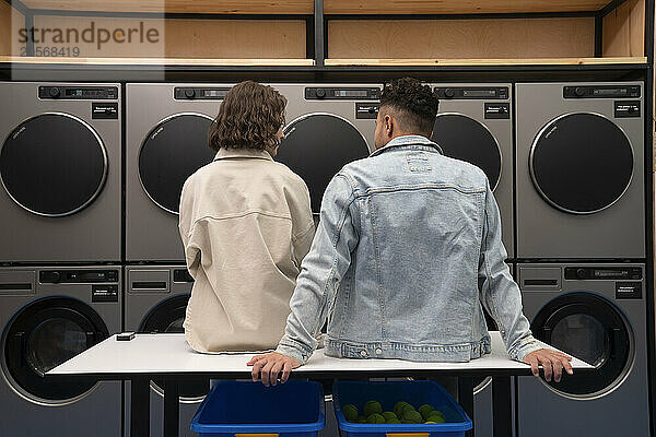 Young boyfriend and girlfriend sitting on table in front of washing machines at laundromat