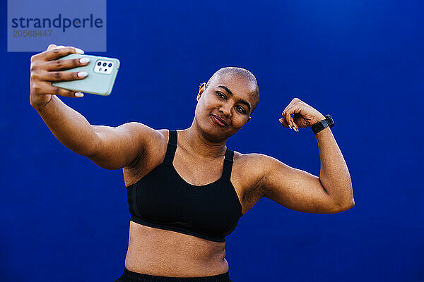 Woman flexing muscles and taking selfie with smart phone in front of blue wall