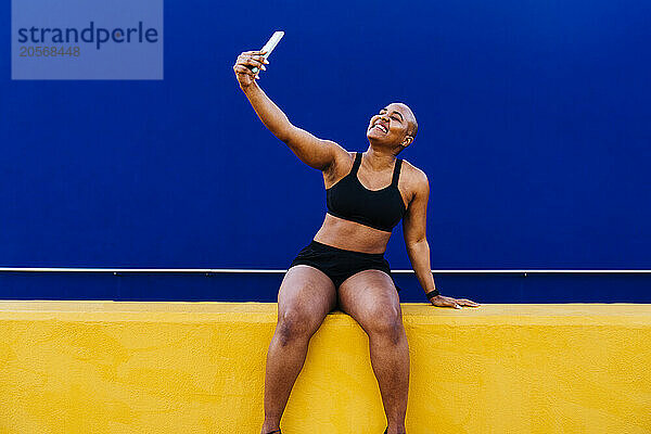 Happy bald woman taking selfie through smart phone in front of blue wall