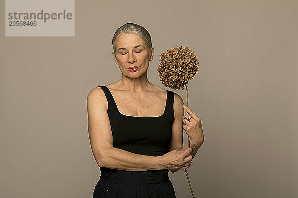 Senior woman with eyes closed holding withered flower in front of brown background