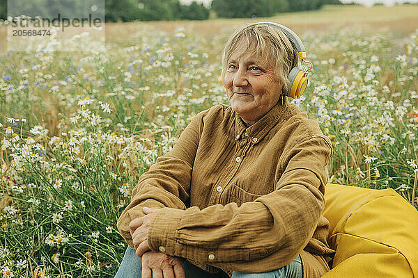 Smiling senior woman wearing wireless headphones and sitting on bean bag at chamomile field