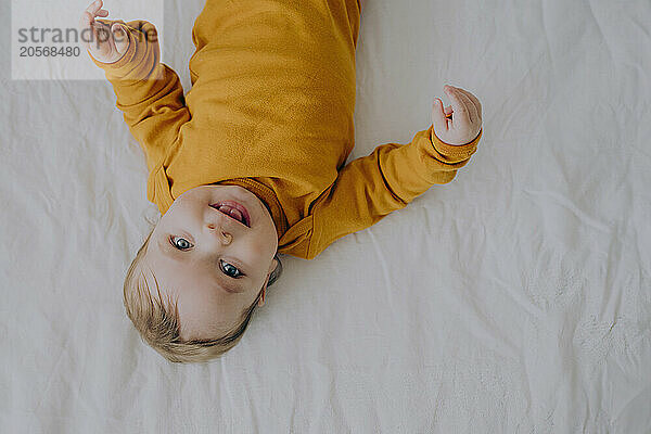 Playful baby boy in orange bodysuit lying on back in bed at home