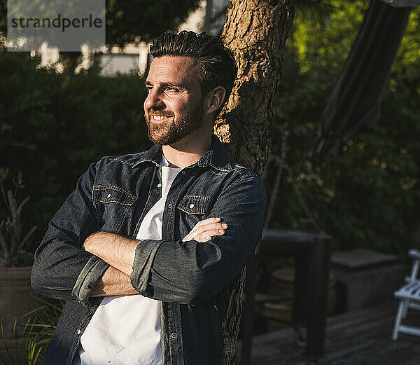 Thoughtful man with arms crossed leaning on tree trunk