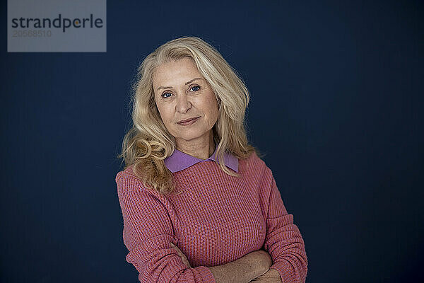 Blond senior woman with arms crossed in front of blue wall