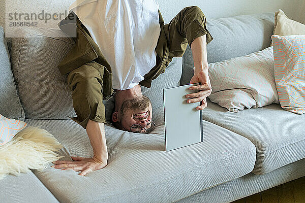 Smiling young active man using tablet PC doing headstand on sofa at home