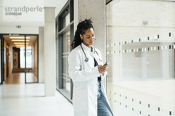 Young female doctor wearing lab coat using smart phone leaning on column in hospital corridor