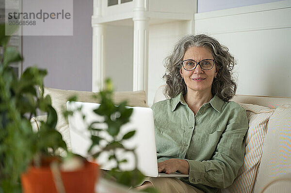 Beautiful gray hair woman in eyeglasses sitting with laptop on sofa at home