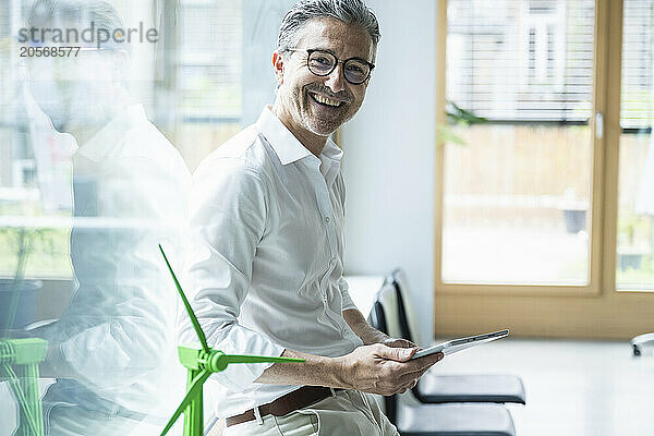 Happy businessman with tablet PC by wind turbine model in office