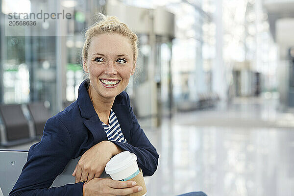 Happy businesswoman holding disposable coffee cup and sitting in airport lobby