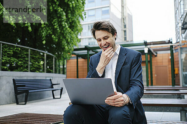 Happy businessman with hand on chin using laptop in office park