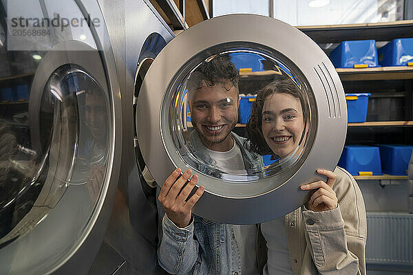 Smiling young couple looking through washing machine glass at laundromat