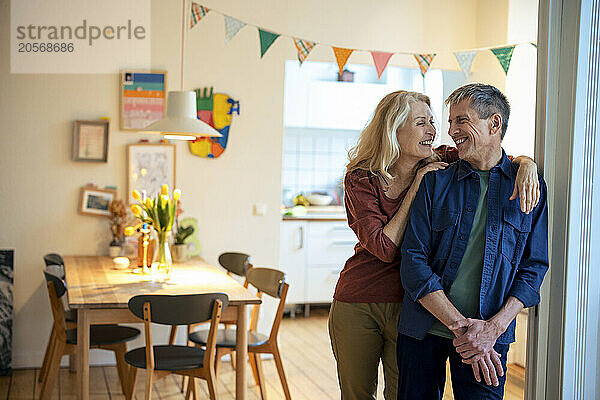 Happy woman with arm around man leaning on wall at home